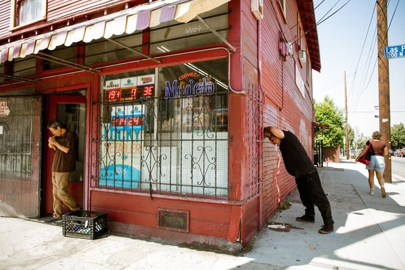 One man lighting a cigarette, wearing a chocolate brown polo shirt and tan pants exists a bodega, while just around the corner another man wearing all black is puking up his guts.