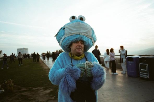 A man standing on a busy promenade wearing a Cookie Monster costume at twilight. Cookie’s head has a surgical mask on its face. It’s cocked back to reveal the man’s actual face with an awkward smile. He has a man purse slung around his neck, a yellow scarf, and a fistful of dollar bills in his hand which he seems to be counting.