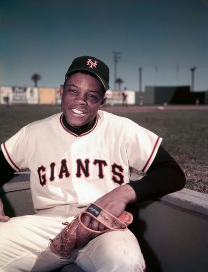A promotional photo in color of Willie Mays sitting in the dugout with his back to the field, wearing his NY Giants uniform, circa 1950s.