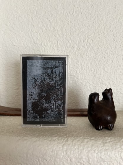A photo of the cassette case for “The Warm Garden” siting on my mantle. The abstract artwork is screen printed in white.