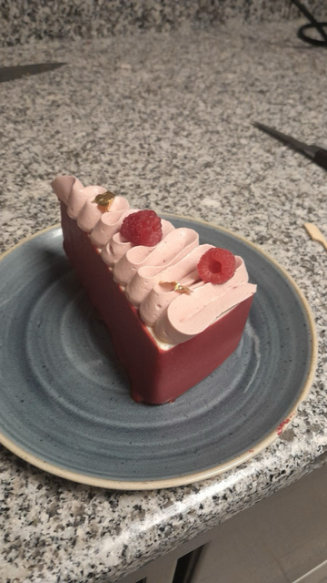 Un très joli gâteau en forme de triangle allongé avec des bords rose foncé et surmonté d’une jolie couche de crème rose pale (comme des froufrous) et 2-3 framboises 