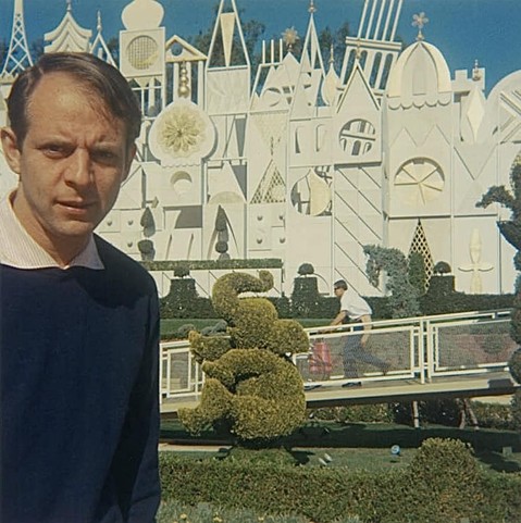 A color photo of a young Karlheinz Stockhausen standing in front of the “It’s a Small World” attraction at Disneyland in Anaheim, California and looking just a bit too serious.