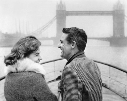 Ingrid Bergman (left) and Carey Grant (right) wearing warm coats while standing on the deck of a boat. In the distance is London Bridge.