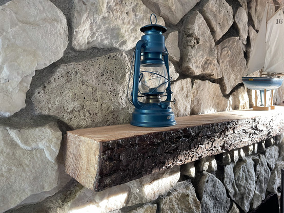A rough-cut slab mantlepiece on a stone fireplace, with an oil hurricane lamp on it.