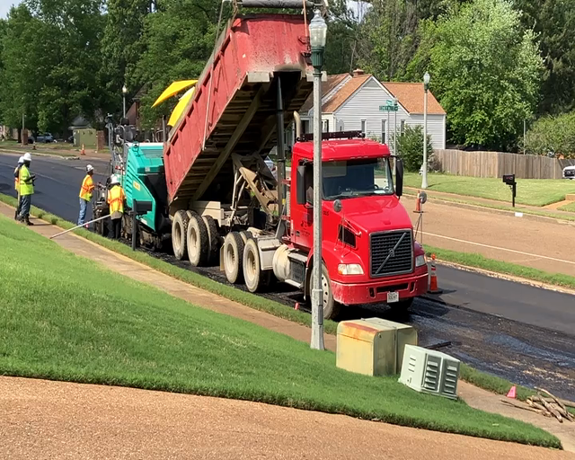 A Lehman-Roberts paving crew is paving the outside portion of a residential street lane. In this video you can see a dump truck using its hydraulic lift to gradually empty its load of asphalt into the hopper of a big Wirtgen / Vögele paving machine, as they both move forward and the paving machine be doin' what a paving machine do.