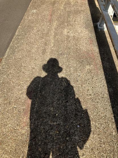 A shadow of a man wearing a floppy hat is cast on some concrete, with the sun behind the photographer