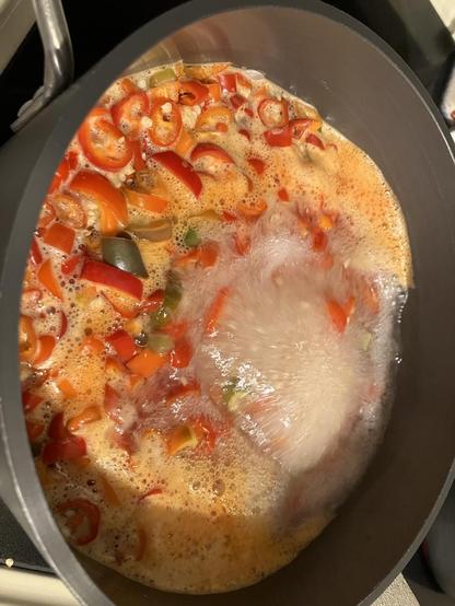 A batch of (mostly) ripe red chile pepper slices are boiling in a pot with water, vinegar, salt, sugar, and a few cloves of garlic.