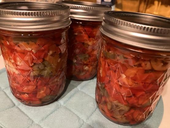 Three jars of freshly canned chile pepper slices are cooling on a pot holder after being sealed. The contents are mostly red in color.