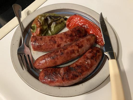 A plate with three grilled bratwurst, plump and brown, with two red peppers and one green pepper 