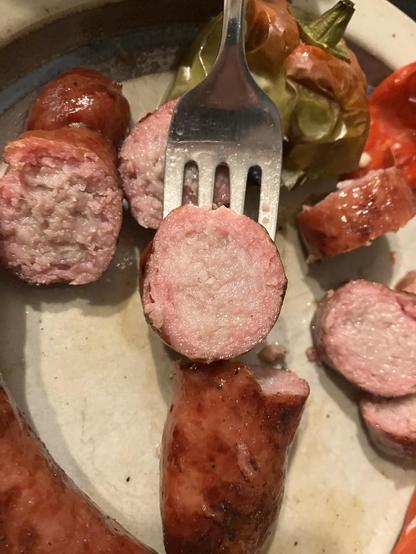 A plate with three grilled bratwurst, plump and brown, with two red peppers and one green pepper. A brat has been sliced open showing a pink smoke ring around the outside 