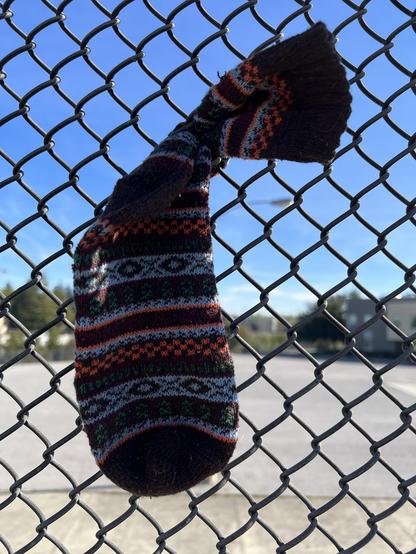 A lone sock with colorful striped patterns hangs from a chain link fence.