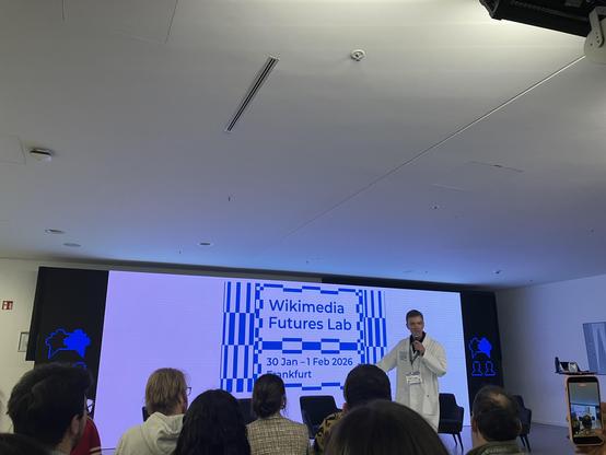 Someone wearing a lab coat is standing in front of a slide that says Wikimedia Futures Lab
