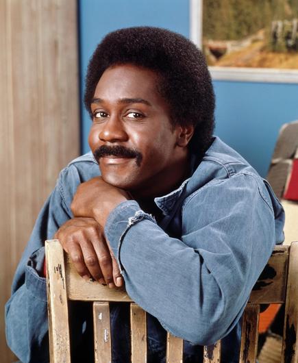 Sanford & Son era photo of Demond leaning on the back of a wooden chair.