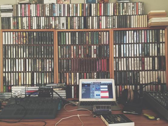 A mixing board connected to a laptop in front of a modest cassette tape library.