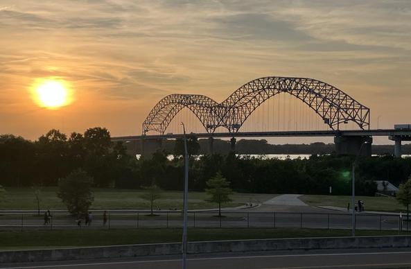 The sun sets over Memphis, Tennessee. The iconic Hernando de Soto Bridge spans the Mississippi River (the river is not visible) as a blazing orange sun falls behind thin clouds.