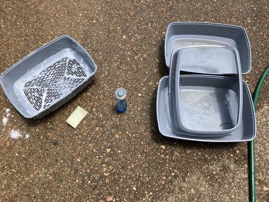 Various components of a sifting cat litter box sit in my driveway. The sifter part is covered in soapy foam and everything is wet.