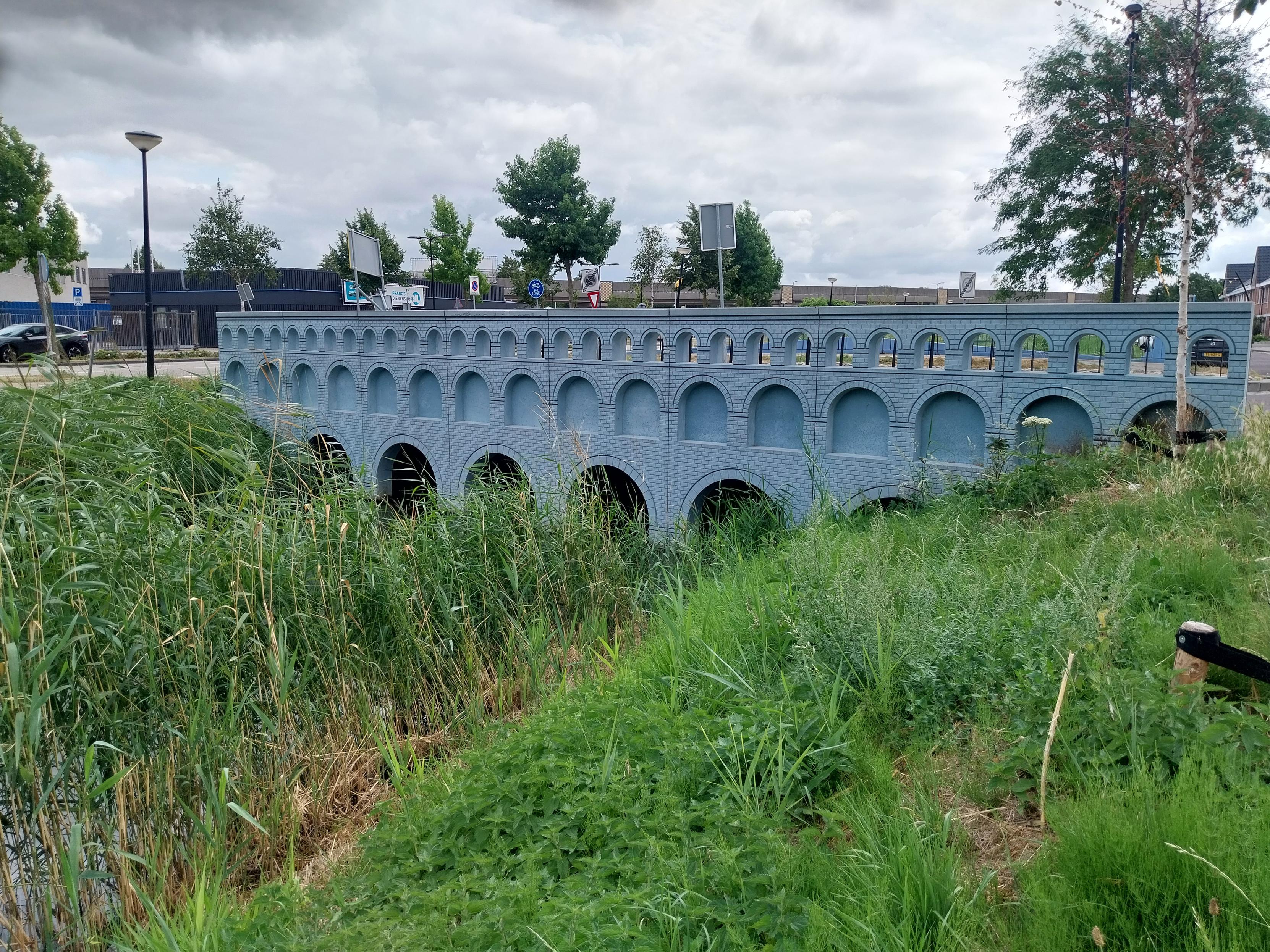 A bridge crossing a small waterway with green banks on either side. The bridge is blue-ish, and designed to look like the bridge on a 5 euro note.
