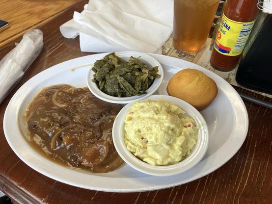 A plate with generous portions of liver & onions, potato salad, turnip greens, and a cornbread muffin. A glass of iced tea with lemonade and a bottle of hot sauce are in the background.
