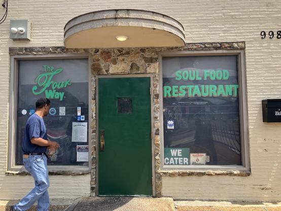 The front of the famous Four Way Soul Food Restaurant at 998 Mississippi Boulevard in Memphis, Tennessee. A green door sits between two large windows. From left of frame, a man walks toward the door. 