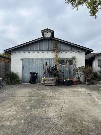 On an overcast day, a somewhat weathered house with faded grey-blue farm style garage door, a garbage can, and a bunch of plants in varying size planters blocking entry into the garage.