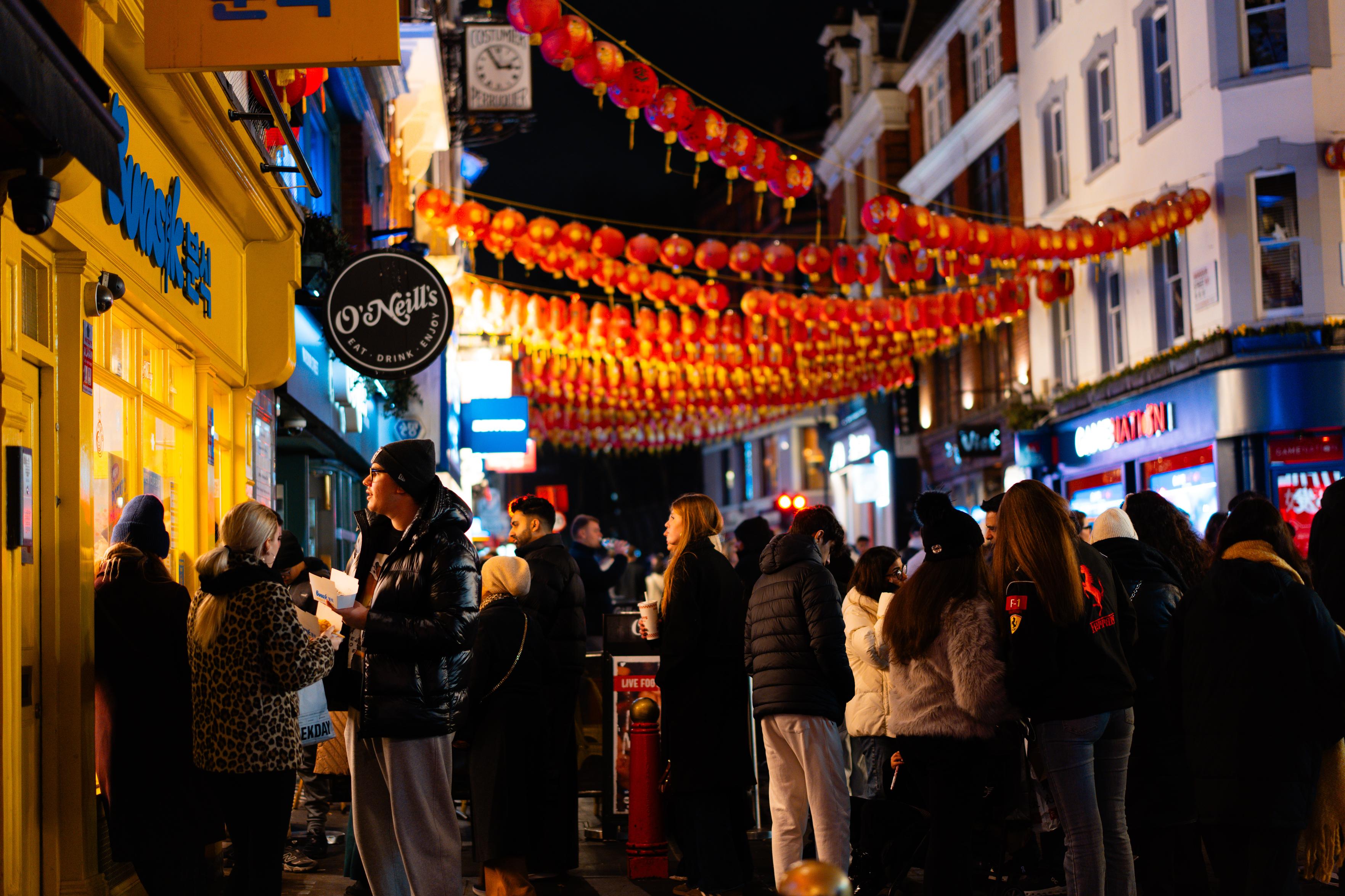 A packed Chinatown street at night, seen at eye level through the crowd. Several long rows of red lanterns stretch overhead between the buildings, receding into the distance. On the left, a bright yellow Korean street-food shop front and a round black O'Neill's pub sign; a hanging clock sign reading "Costumier Perruquier" is above. People in puffer jackets and beanies stand around eating from paper cartons and drinking from takeaway cups.