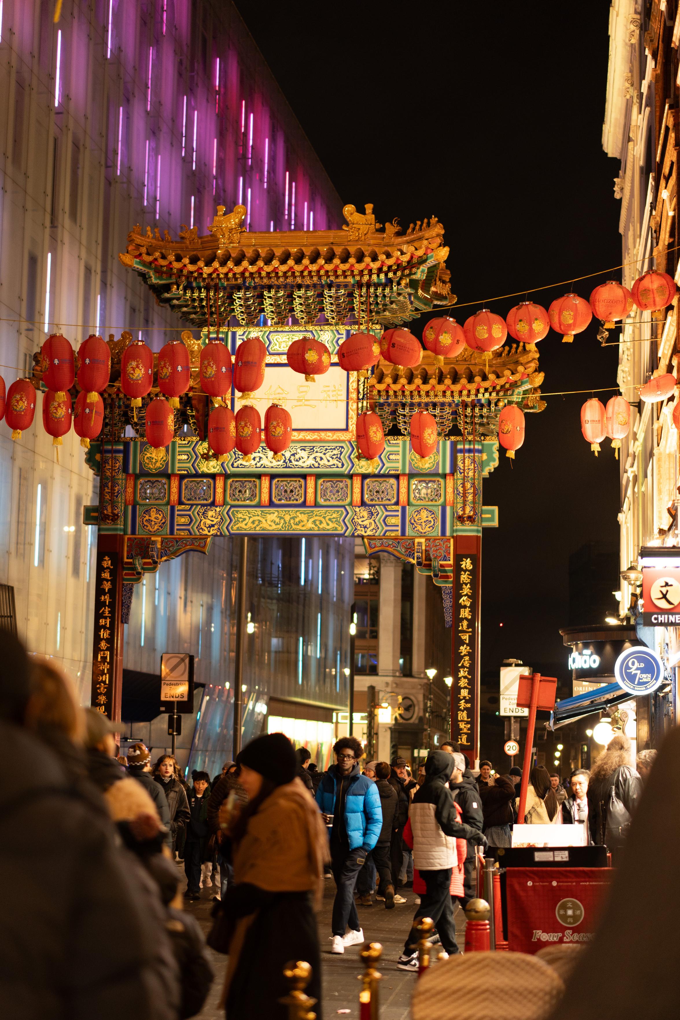 London's Chinatown Gate at night — an ornate paifang painted in red, green, gold and blue, with a tiled double roof, carved dragon panels and gilded figures along the ridge. Dozens of red lanterns hang from wires strung across the street in front of it, and a crowd of people in winter coats walks beneath. A large building on the left is lit with vertical strips of purple and pink light.