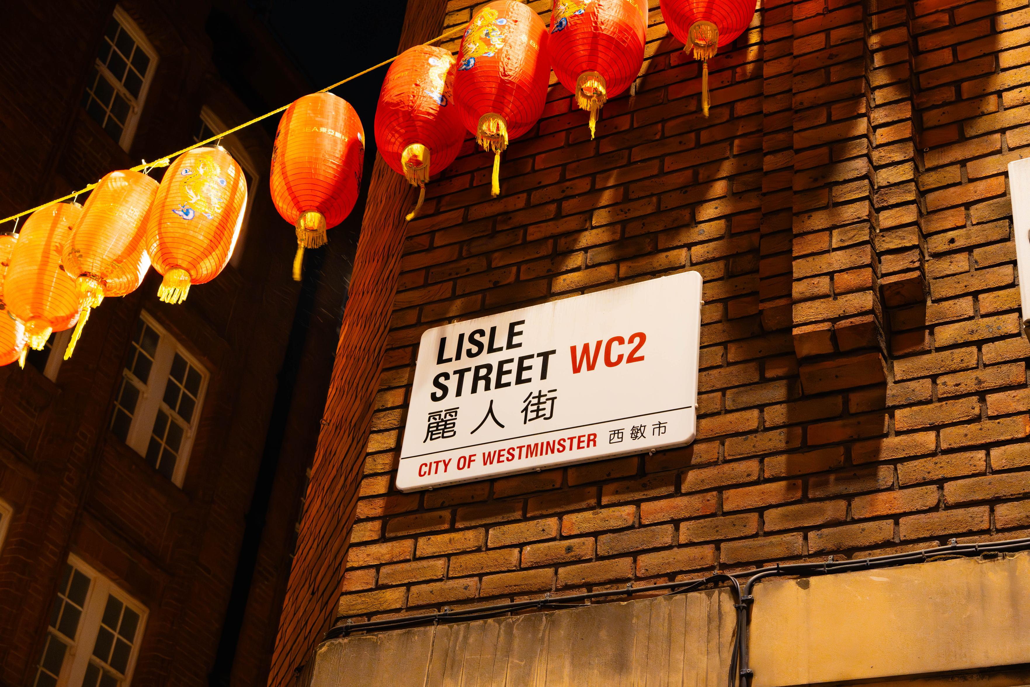Looking up at a white City of Westminster street sign reading "LISLE STREET WC2" with the Chinese characters 麗人街 beneath, mounted on a warmly lit red-brick corner. A string of glowing red-and-orange paper lanterns with gold tassels runs diagonally across the dark night sky above it.