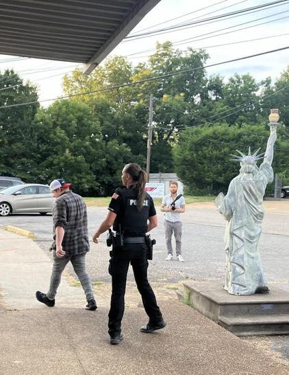 Outside the police station in Mason, Tennessee, where the town’s aldermen were having a meeting. A resident has walked out of the meeting in frustration, and exited the building to commiserate with others outside. For reasons that aren’t clear, a police officer is hustling outside after him to run him off the property, where he ought to have every legal right to be. Watching the whole scene unfold is a replica of the Statue of Liberty, which stands on a pedestal outside of the police station. A…