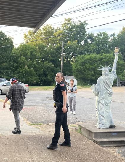 Outside the police station in Mason, Tennessee, where the town’s aldermen were having a meeting. A resident has walked out of the meeting in frustration, and exited the building to commiserate with others outside. For reasons that aren’t clear, a police officer is hustling outside after him to run him off the property, where he ought to have every legal right to be. Having chased him away, she now pivots and looks at me taking her picture. Watching the whole scene unfold is a replica of the Sta…