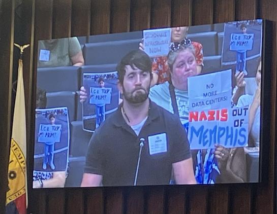Memphis City Council chambers, September 1, 2026. As a resident gives a public comment, audience members seated behind him hold signs for the camera: 

NAZIS OUT OF MEMPHIS
ICE TOOK MY MOM
NO MORE DATA CENTERS
SUSTAINABLE INFRASTRUCTURE NOW