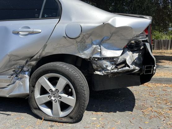 Close-up view of the carnage. Left rear wheel is pushed in at an angle and a part of the rear quarter panel is missing to reveal the car’s guts.