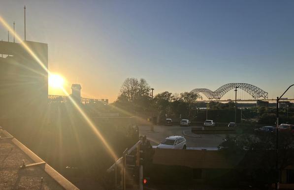 Facing west, the sun is setting directly into the camera in downtown Memphis, Tennessee. In the foreground, parts of Mud Island and its parking garage are visible. Behind is the Hernando de Soto Bridge against the horizon.